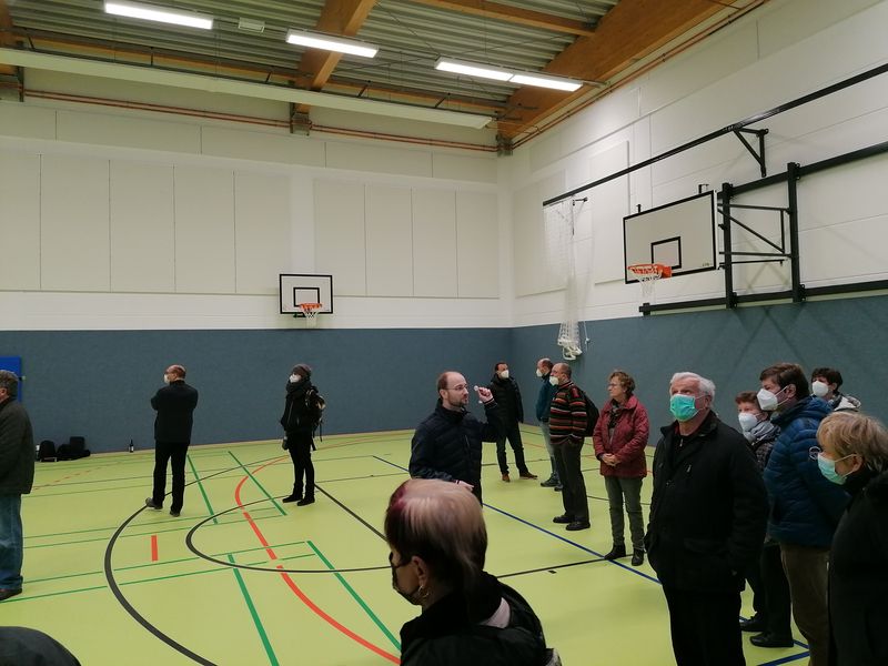 Eine Gruppe von Menschen mit Masken steht verteilt in einer modernen, hellen Sporthalle.