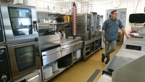 Es zeigt eine modern ausgestattete, professionelle Großküche. Im Mittelpunkt steht ein Mann, der lässig an einer Arbeitsplatte lehnt.