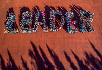Eine Gruppe von Menschen formt in kleineren Gruppen die Buchstaben des Wortes LEADER. Die Szene ist aus der Draufsicht auf einem roten Sportplatz aufgenommen, am unteren Bildrand sind Schatten von Bäumen zu sehen.