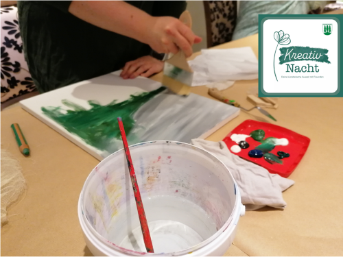 Person malt mit breitem Pinsel auf Leinwand; Utensilien und Logo „Kreativ Nacht“ sind sichtbar.