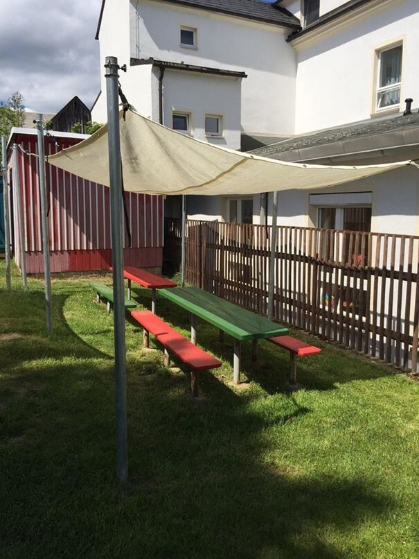 Bunte Sitzbänke und Tische unter einem Sonnensegel auf einer Rasenfläche