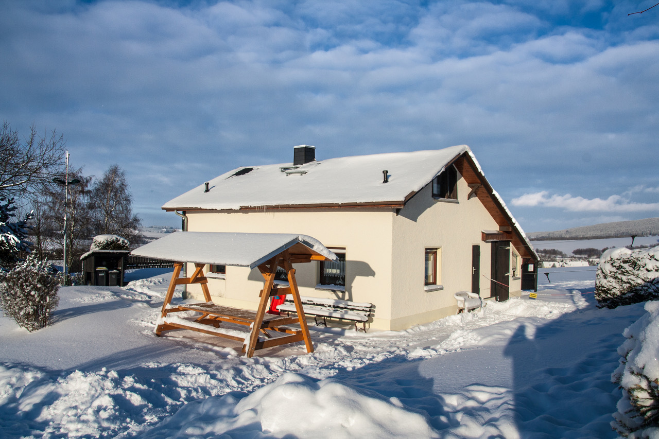 Verschneite Skihütte mit überdachter Sitzgruppe in einer Winterlandschaft. (Link öffnet Bild vergrößert in einem Popupfenster)