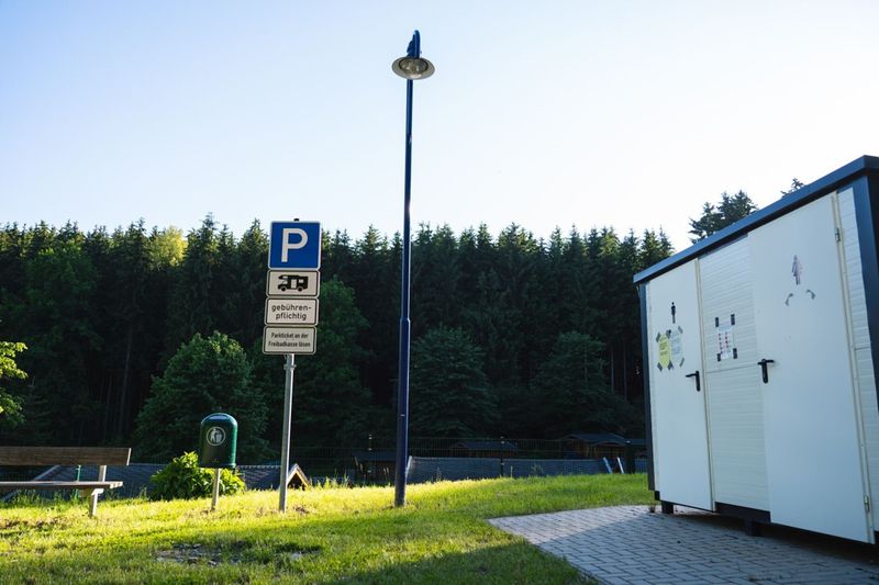 Query successful  Ein markiertes Parkplatzschild für Wohnmobile vor einem dichten Waldpanorama und einer Sanitäranlage.  (Link öffnet das Foto vergrößert in einem Popupfenster)