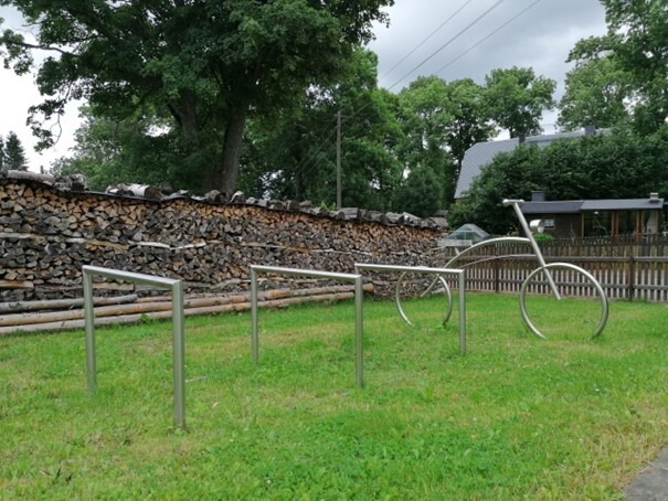 Query successful Drei metallene Fahrradanlehnbügel auf einer Wiese vor einer hohen Holzstapelwand und einer großen Fahrradskulptur. (Link öffnet Bild vergrößert in einem Popupfenster).