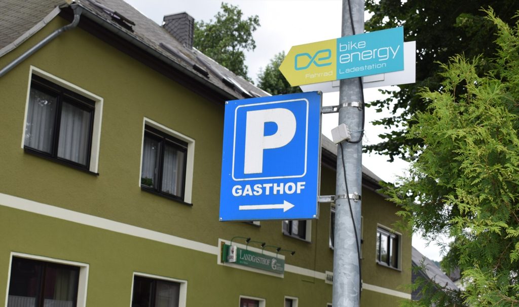 Query successful Blaues Parkplatzschild für einen Gasthof mit einem Hinweis auf eine „bike energy“ E-Bike-Ladestation direkt darüber. (Link öffnet Bild vergrößert in einem Popupfenster).