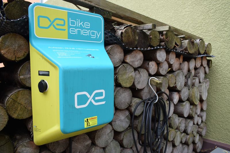 Eine blau-gelbe „bike energy“ E-Bike-Ladestation, montiert an einem Holzstapel, mit einem daneben hängenden Ladekabel. (Link öffnet Bild vergrößert in einem Popupfenster).