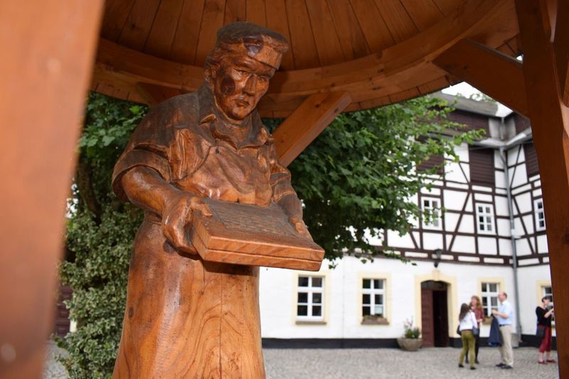 Geschnitzte Holzfigur eines Papiermachers mit Schöpfsieb vor einem weißen Fachwerkhaus. (Der Link öffnet Bild vergrößert in einem Popupfenster)