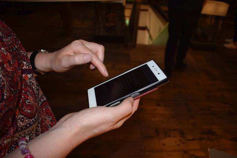 Nahaufnahme von Händen, die ein Smartphone vor einem rustikalen Holzboden bedienen. (Link öffnet Bild vergrößert in einem Popupfenster).