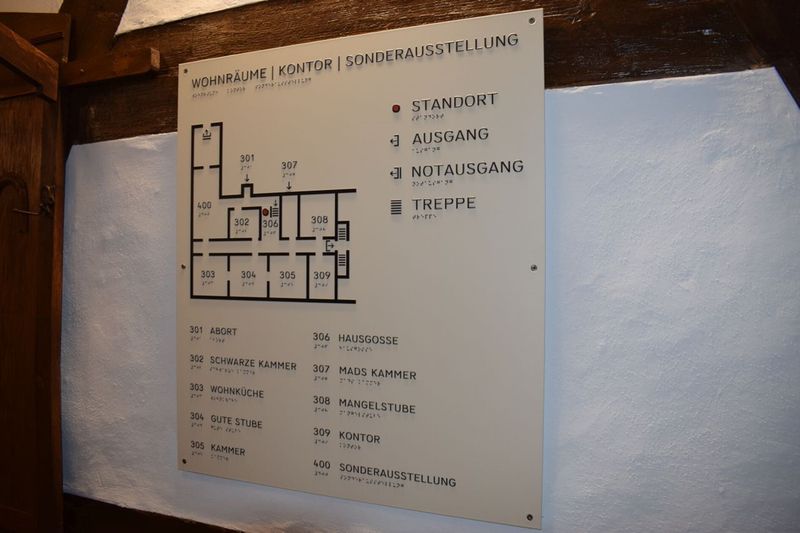 Weißes Hinweisschild mit Lageplan, Raumnummern und Brailleschrift an einer rustikalen Wand. (Link öffnet Bild vergrößert in einem Popupfenster).