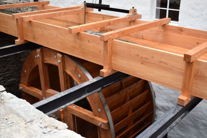 Hölzernes Wasserrad mit Zuleitungsrinne im Außenbereich vor einer weißen Steinwand. (Link öffnet Bild vergrößert in einem Popupfenster).