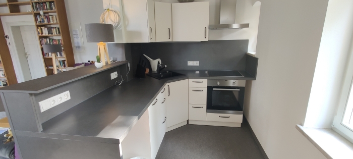 Moderne, helle Eckküche mit dunkler Arbeitsplatte, weißen Fronten, Herd, Spüle und Bücherregal im Hintergrund. (Link des Bildes öffnet sich in vergrößerter Darstellung als Popup-Fenster)