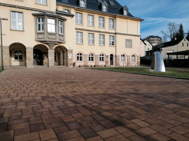 Gepflasterter Vorplatz vor einem großen gelben Gebäude mit Erker, Rundbögen und moderner Skulptur. (Link öffnet Bild vergrößert in einem Popupfenster)