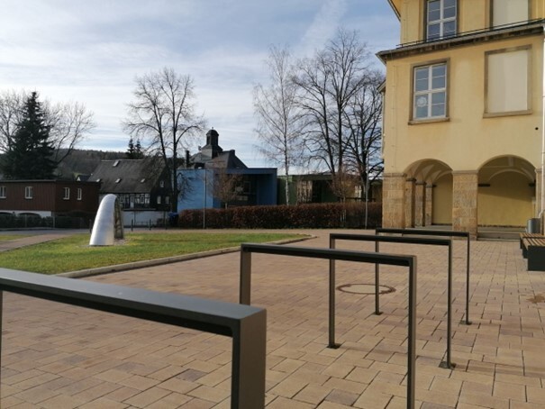 Gepflasterter Vorplatz mit Fahrradständern vor einem gelben Gebäude (die Oberschule) und moderner Skulptur links. (Link öffnet Bild vergrößert in einem Popupfenster)