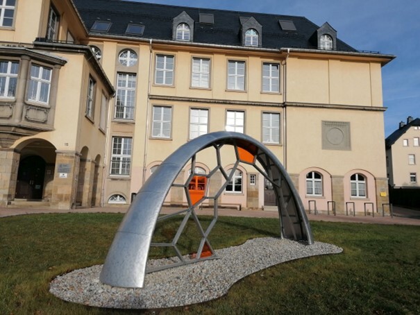 Bogenförmige Skulptur aus Metall mit orangefarbenen Glaselementen auf einer Rasenfläche. (Link öffnet Bild vergrößert in einem Popupfenster)