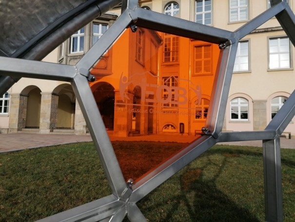 Nahaufnahme einer orangefarbenen Glasscheibe in einer Metallskulptur mit dem Logo „EOB“ - im Hintergrund sieht man das Schulgebäude. (Link öffnet Bild vergrößert in einem Popupfenster)