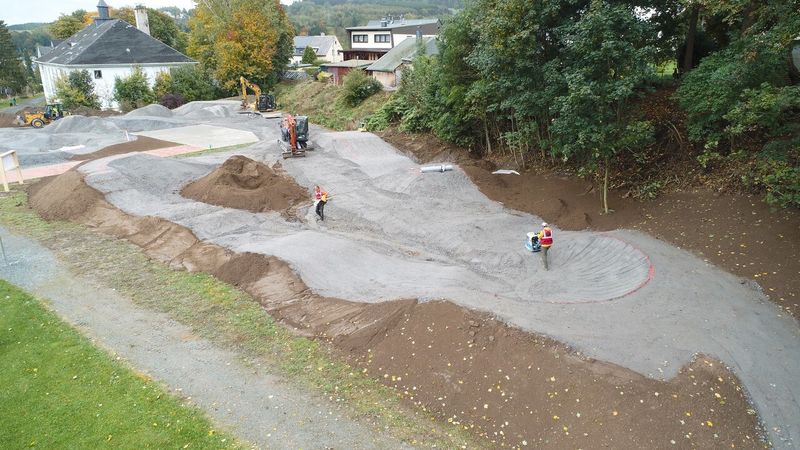 Luftaufnahme eines im Bau befindlichen Pumptracks mit Hügeln aus grauem Schotter und Erde sowie Bauarbeitern. (Link öffnet Bild vergrößert in einem Popupfenster).