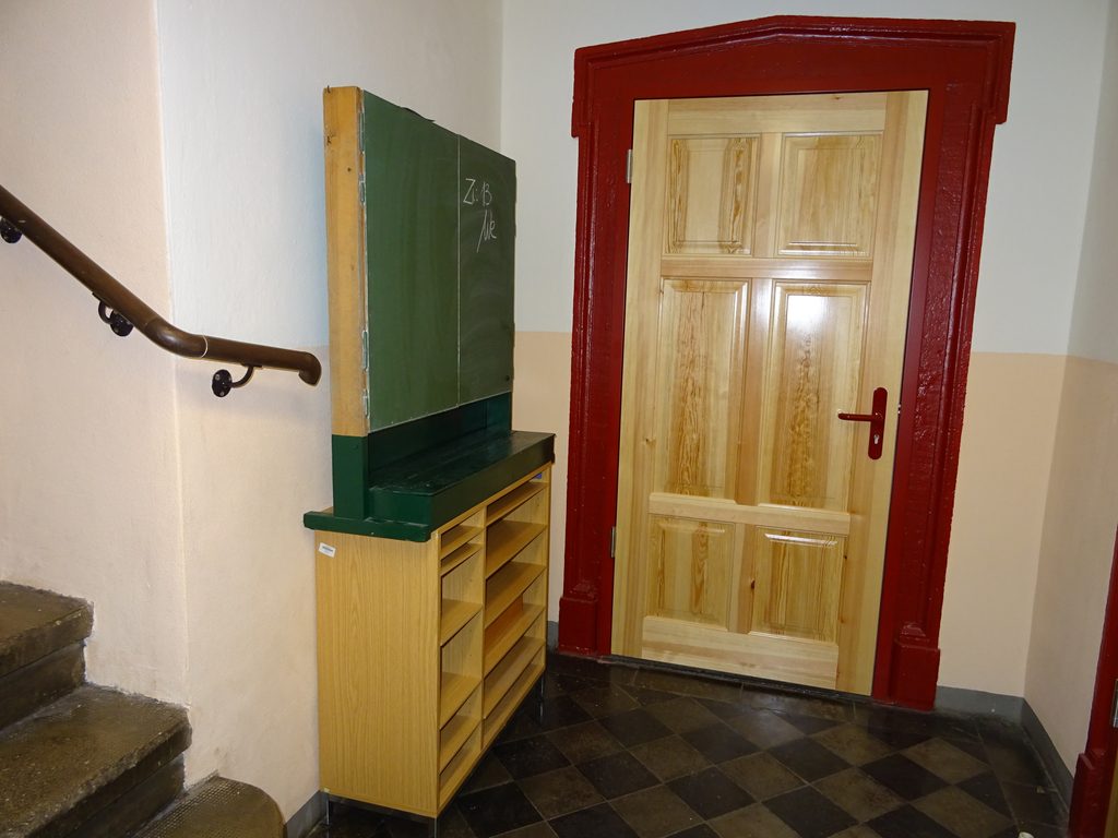 Eine hellbraune Holztür auf einem roten Türrahmen. Links daneben befindet sich ein Regalschrank und davor geht links eine Steintreppe hoch.