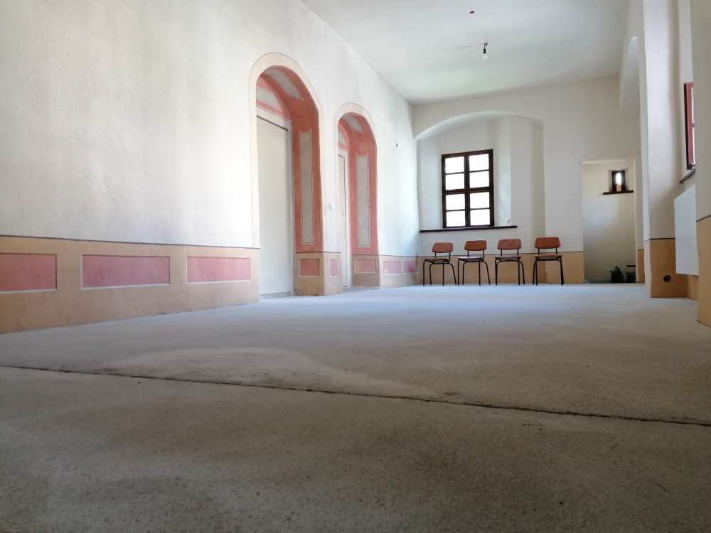 Großer, heller Raum im Rohbauzustand mit Rundbögen, rosa Wandakzenten und einer kleinen Stuhlreihe. (Link öffnet das Foto vergrößert in einem Popupfenster)