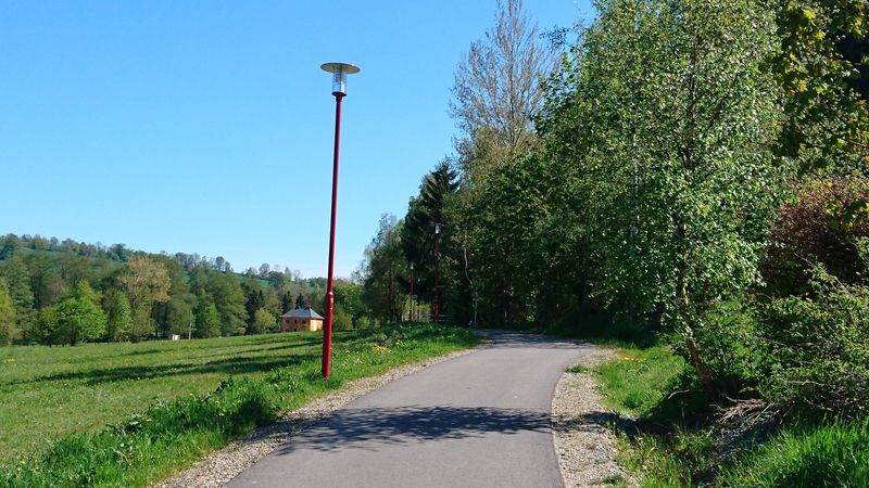 Asphaltierter Weg mit roter Laterne führt durch grüne Wiesen und Bäume