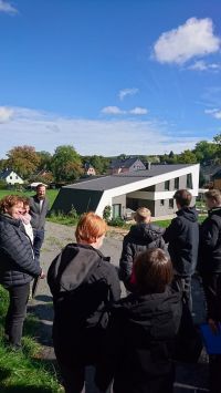 Gruppe von Menschen betrachtet ein modernes Gebäude mit asymmetrischem Dach unter blauem Himmel.