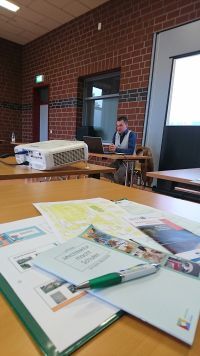 Informationsmaterialien auf einem Tisch im Vordergrund; im Hintergrund hält ein Mann einen Vortrag.