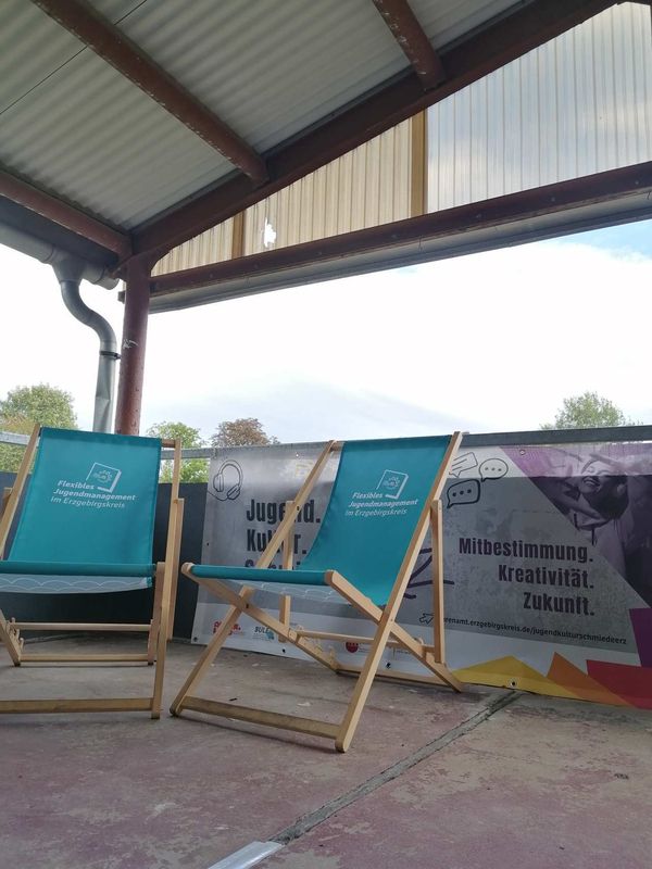 Zwei Liegestühle mit Logo unter einem Vordach vor einem Banner zum Thema Jugend Kultur Schmiede Erzgebirge