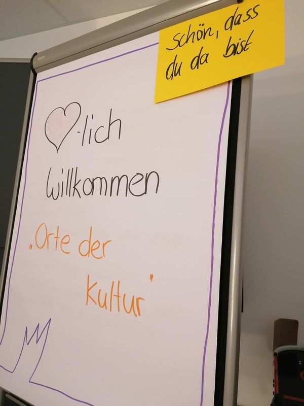 Flipchart mit „Herzlich Willkommen“ und „Orte der Kultur“, oben gelber Zettel: „Schön, dass du da bist“.